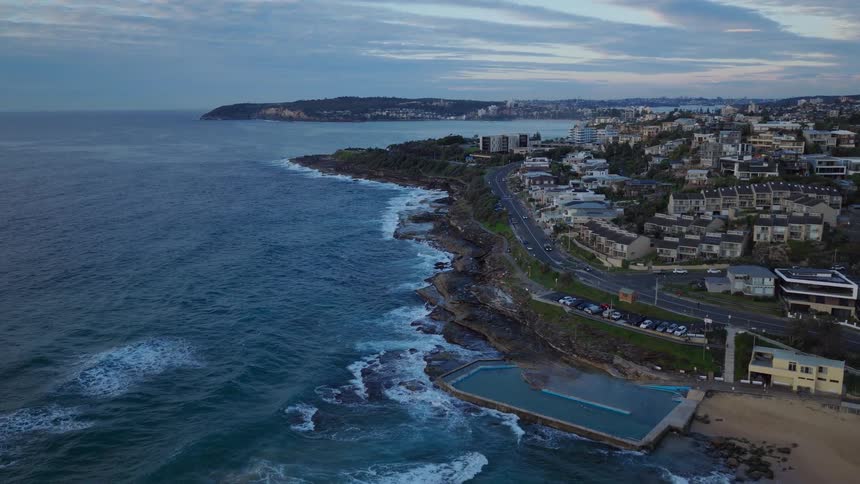A slow aerial pullback reveals the Queenscliff Beach rock pool as waves crash along the rocky coastline of Sydney, Australia at dawn
