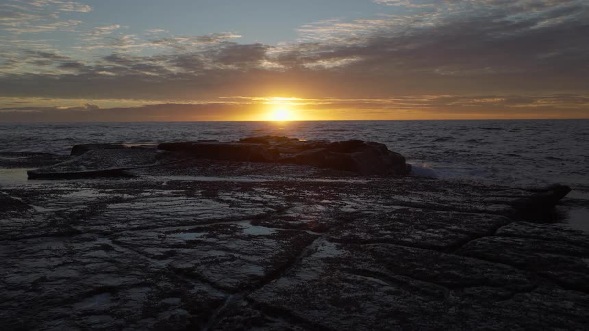 A serene sunrise reflects on the ocean as waves wash over a dark, rocky coastline in the foreground