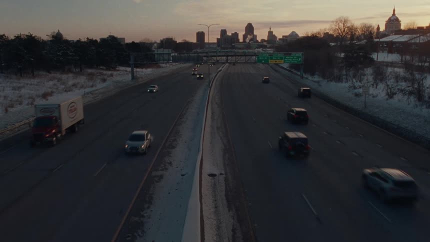 Traffic moves along a snowy highway leading towards a city skyline at sunset during the winter