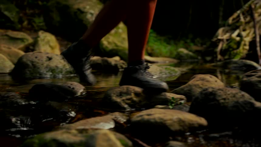A close-up of hikers' feet in boots as they carefully step across mossy rocks in a shallow forest stream