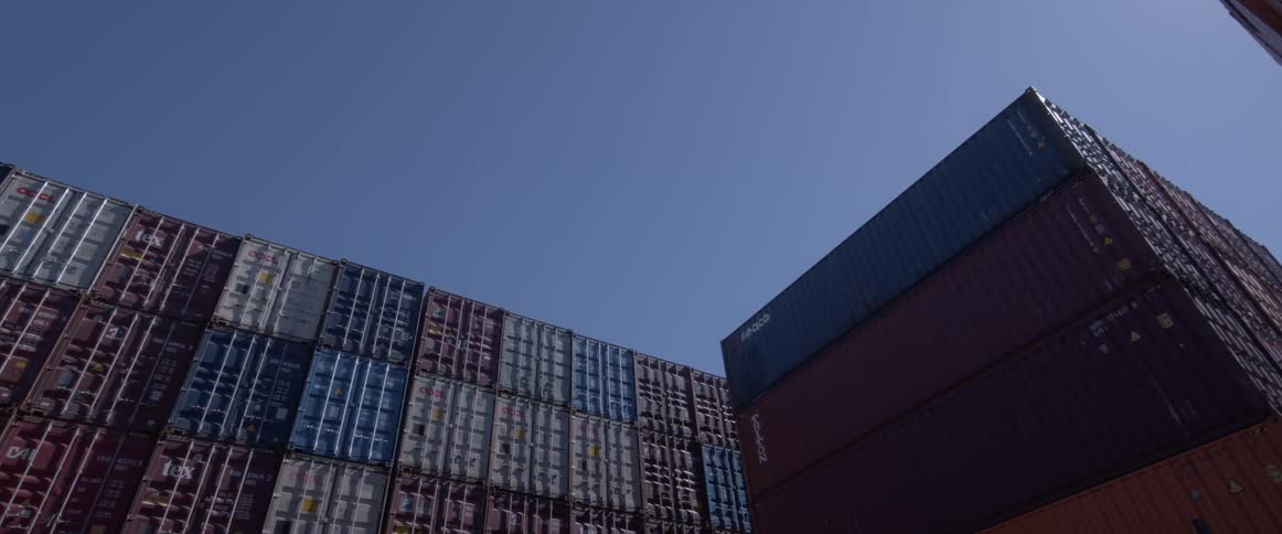 A slow, low-angle pan reveals massive stacks of colorful shipping containers against a clear blue sky at an industrial port