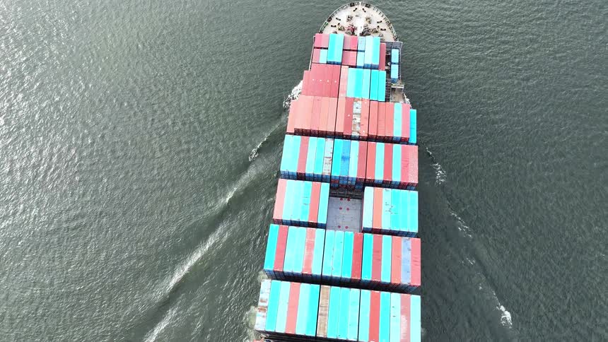 A top-down aerial view captures a large container ship fully loaded with colorful cargo as it moves steadily across the open water