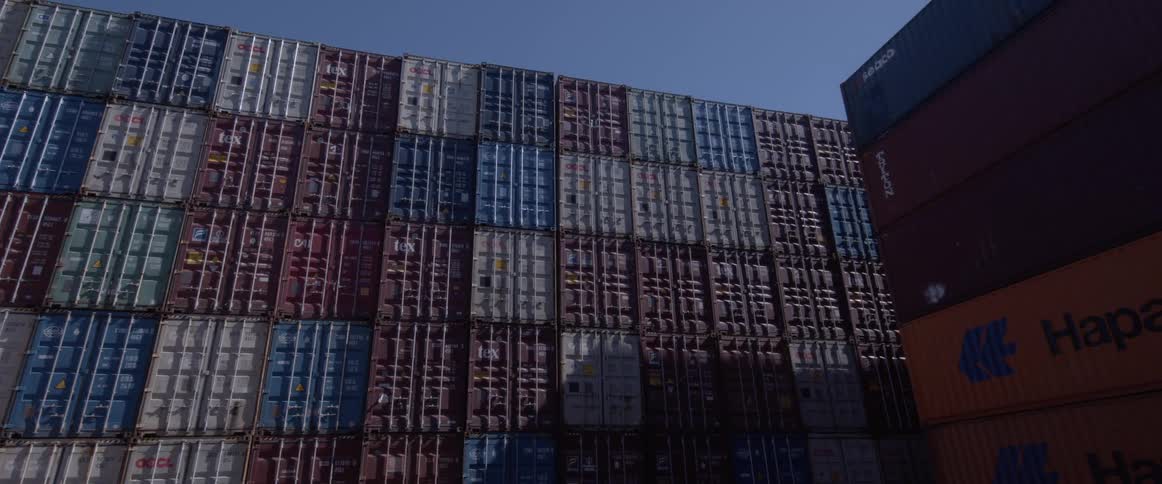 A low angle panning shot reveals a massive wall of colorful shipping containers stacked high in a port