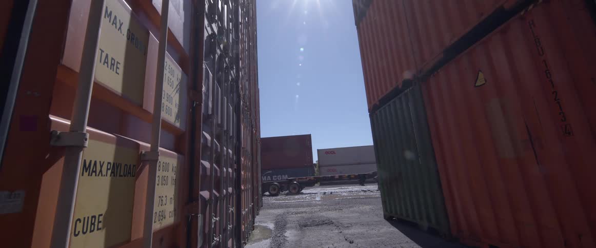 A semi-truck drives away with an empty chassis from between two stacks of shipping containers in a sunny industrial yard
