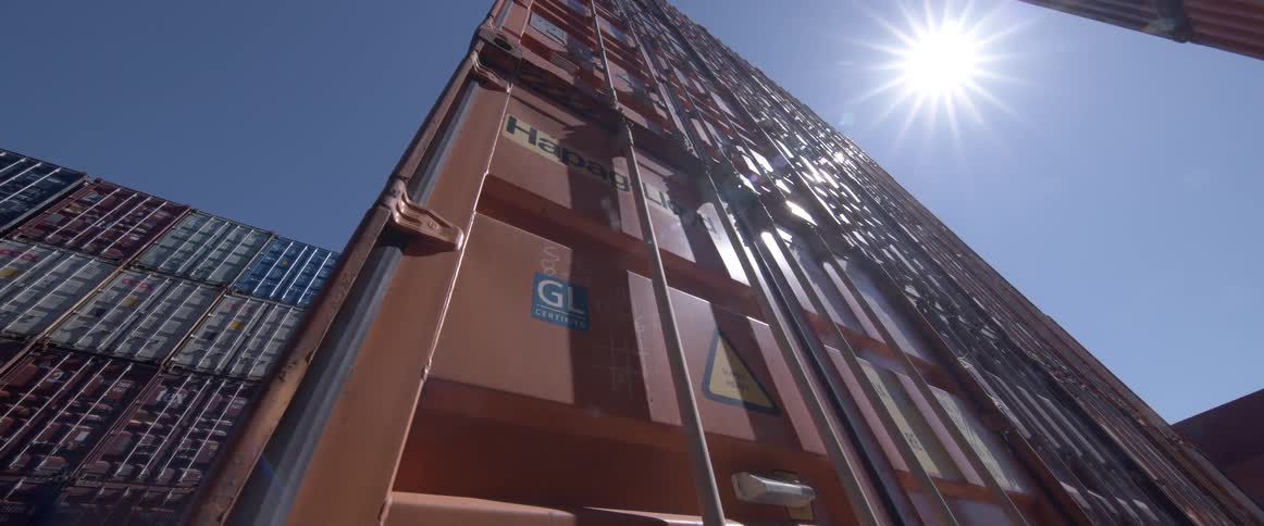 A low-angle shot slowly tilts down the side of a tall orange shipping container in a port on a sunny day