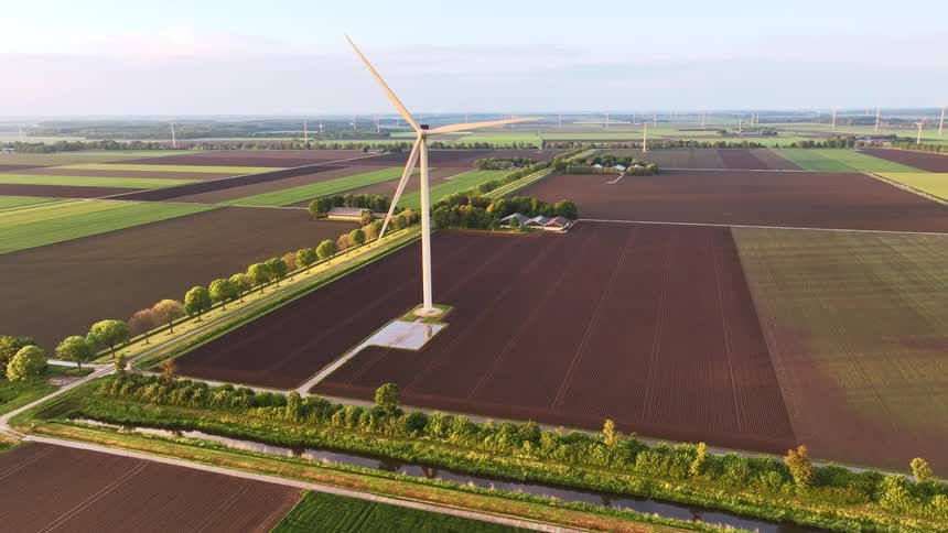A slow aerial pullback reveals a solitary wind turbine amidst a vast patchwork of Dutch farmland at golden hour