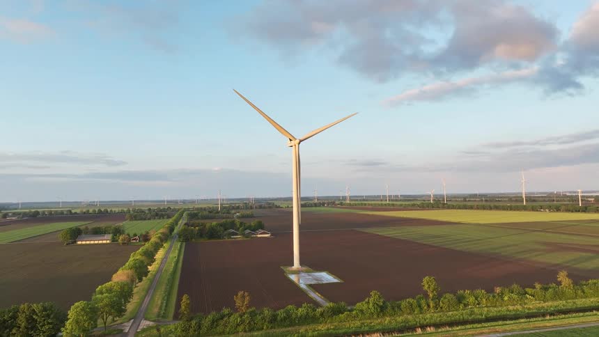 A serene aerial shot captures a large wind turbine turning gracefully amidst expansive farmlands as the sun sets on the horizon