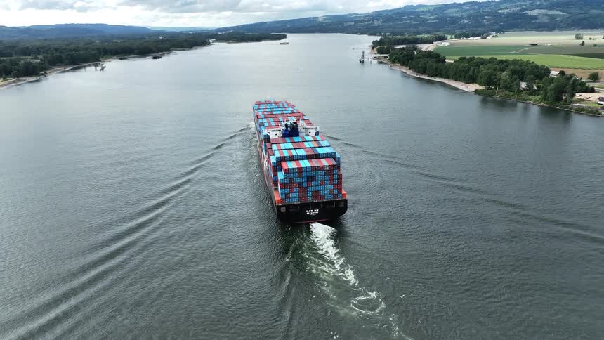 An aerial view from behind follows a fully loaded container ship as it sails down a wide river on a cloudy day