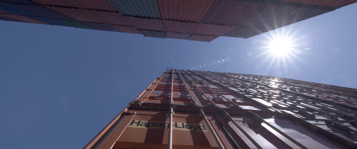 A low angle shot looks up between two tall stacks of orange shipping containers against a bright sun and blue sky