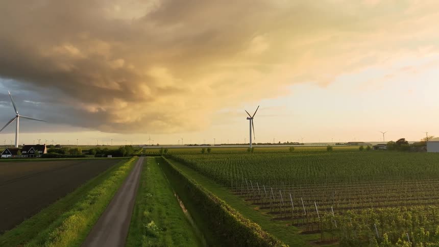 A scenic aerial view shows wind turbines spinning over lush green farmland under a dramatic golden sunset sky