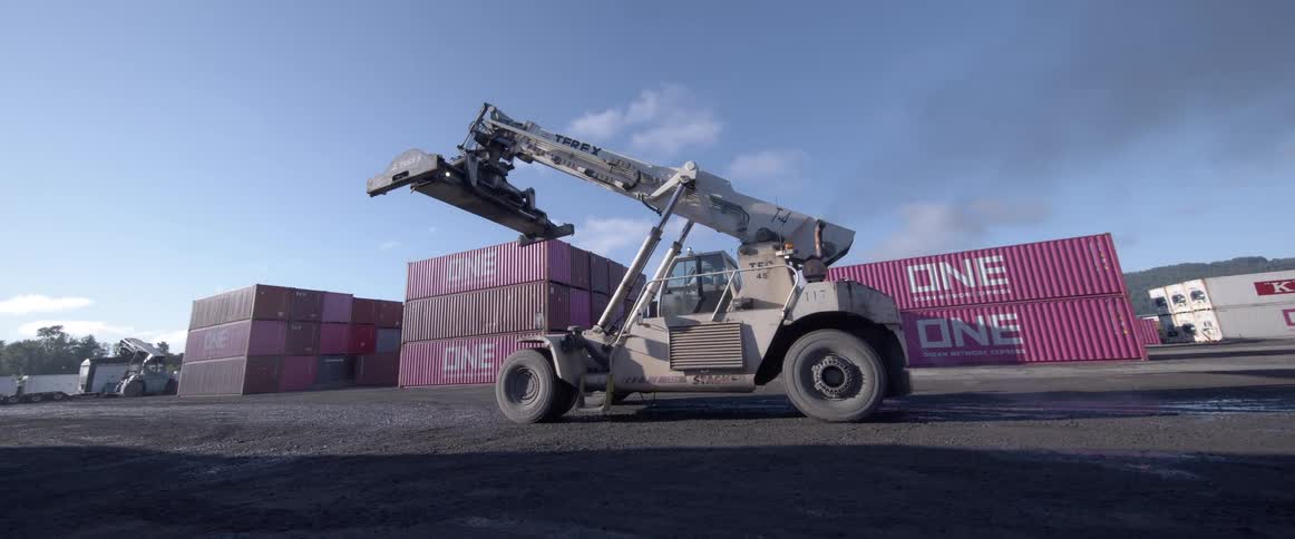 A large industrial reach stacker starts its engine and reverses in a shipping container yard as the camera slowly pulls back