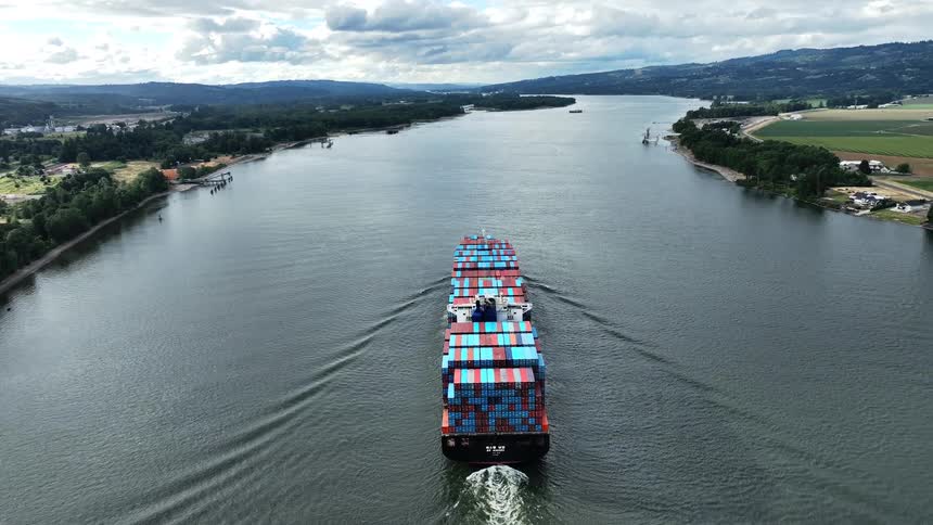 An aerial drone follows a massive cargo freighter fully loaded with colorful containers as it sails down a wide river