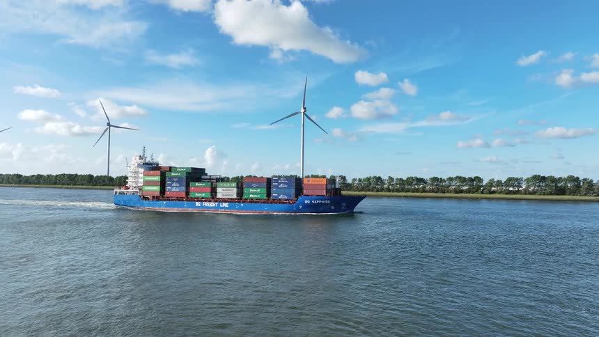 A large container ship sails down a wide canal past spinning wind turbines on a bright, sunny day