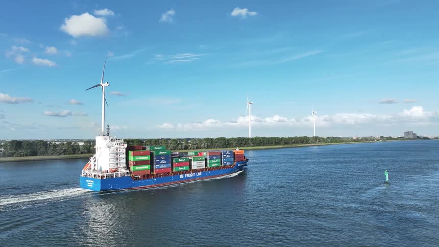 Aerial view of a large container ship sailing down a wide canal past a line of spinning wind turbines under a blue sky