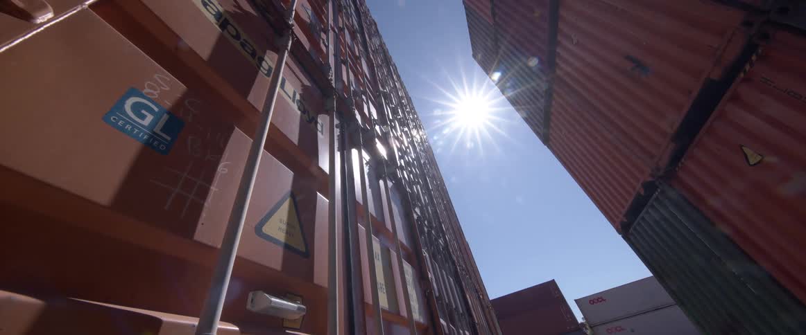 A low angle shot looks up between tall stacks of orange shipping containers as the sun flares brightly in a clear blue sky