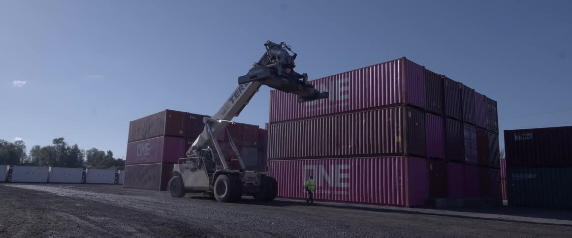 A worker in a high-visibility vest walks past a large reach stacker in a busy shipping container yard on a sunny day