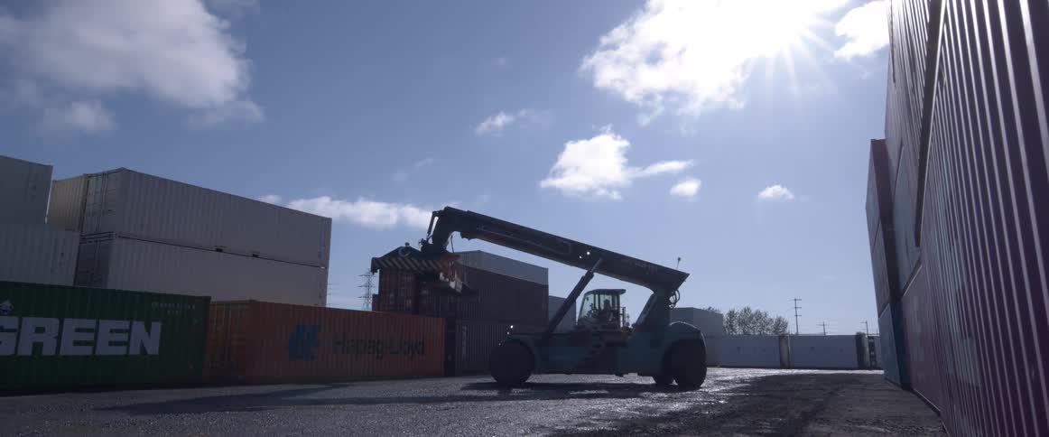 A low-angle view of a large reach stacker preparing to move shipping containers in a port yard against the bright sun