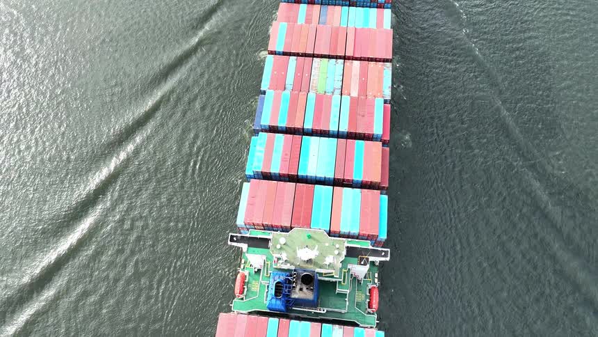 A top-down aerial view follows a massive container ship as it sails through the water, revealing stacks of colorful cargo containers