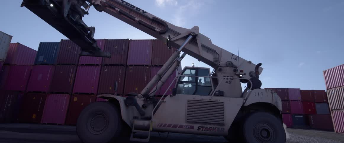 An industrial reach stacker forklift reverses and retracts its boom amidst stacks of colorful shipping containers in a sunny port depot