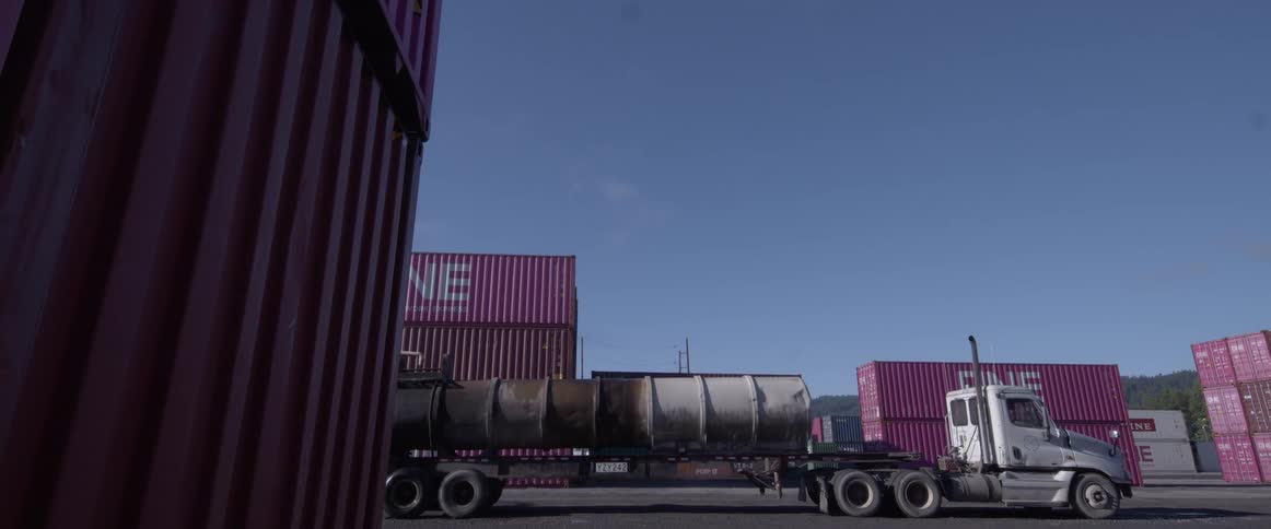 A water tanker truck sprays the ground at a shipping port surrounded by colorful containers on a sunny day