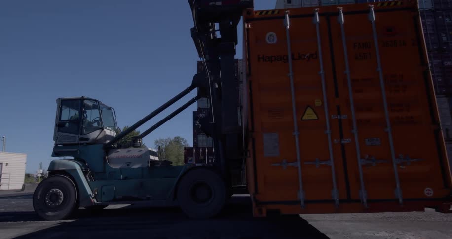 A large container handler forklift slowly backs up, moving an orange shipping container across an industrial depot on a sunny day