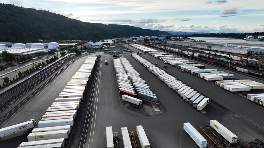 A vast industrial train yard and logistics hub is seen from above, filled with rows of shipping containers and semi-trucks