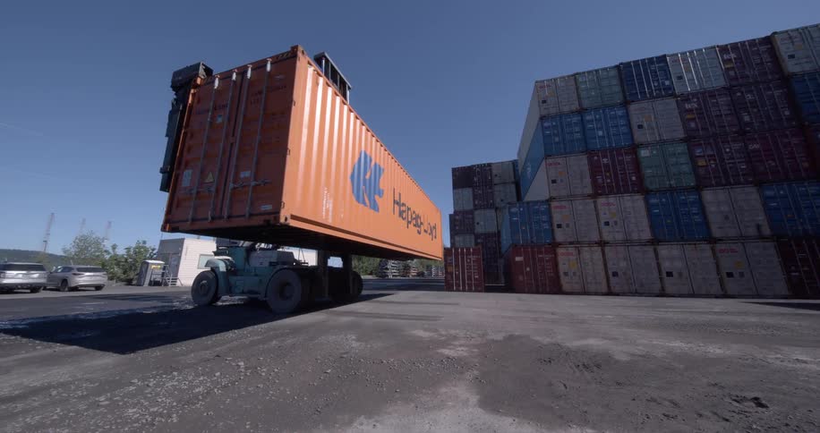 An impressive wide-angle shot shows a reach stacker lifting an orange shipping container and backing up in a large industrial yard