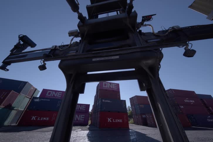 Point of view from a container handler as it reverses through a shipping yard filled with stacked containers under a clear blue sky