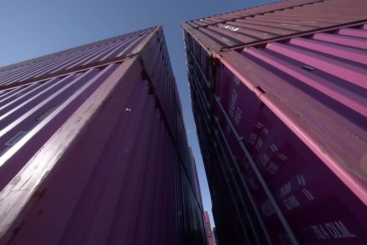 A slow push-in shot from a low angle moves between two tall stacks of purple shipping containers against a clear blue sky