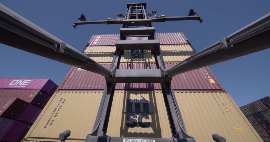 A point-of-view from a container handler forklift as its arms lift towards a tall stack of shipping containers in a port yard