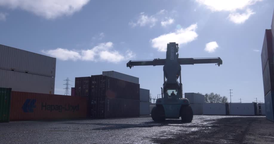 A large reach stacker positions its spreader to lift a shipping container in a depot yard against a bright, sunny sky
