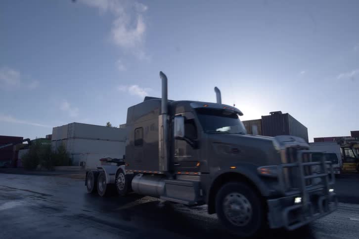 A semi-truck slowly drives through an industrial shipping container yard at sunrise