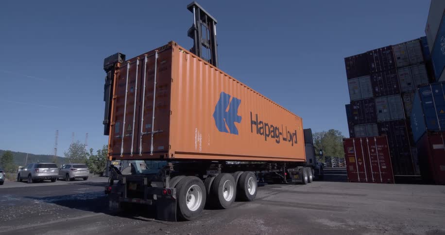 An industrial forklift places a large orange Hapag-Lloyd shipping container onto a semi-truck in a container yard on a sunny day
