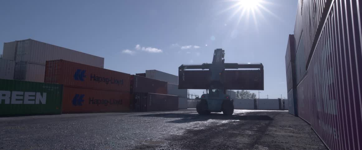 A large container handler lifts and stacks a shipping container in an industrial yard under the bright sun