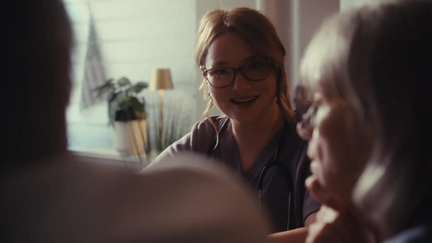 A kind female doctor with glasses consults with an elderly couple in a soft, warmly lit room