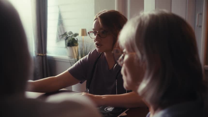 A compassionate nurse consults with an elderly couple in their home, seen from an intimate over-the-shoulder perspective