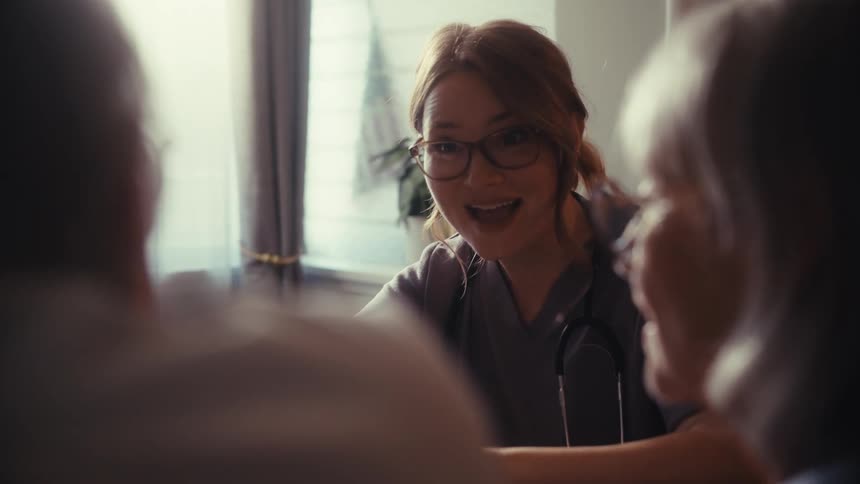A compassionate young female doctor in scrubs consults with an elderly couple during a home visit, offering them reassurance and care