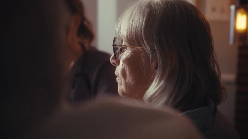 A pensive senior woman with gray hair listens carefully as a female nurse speaks to her during a home visit