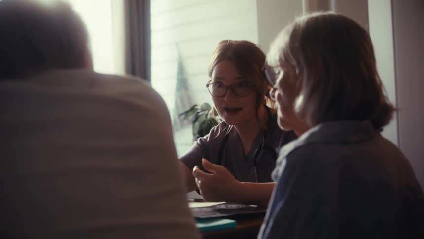 A compassionate female doctor in scrubs consults with a senior couple during a home visit, lit by soft window light