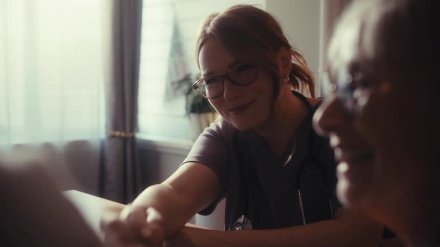 A compassionate young female nurse laughs and holds hands with an elderly couple during a caring home visit