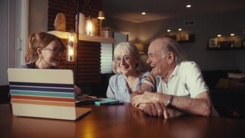 An adult daughter and her elderly parents laugh heartily while sitting at a dining table looking at a laptop and photos