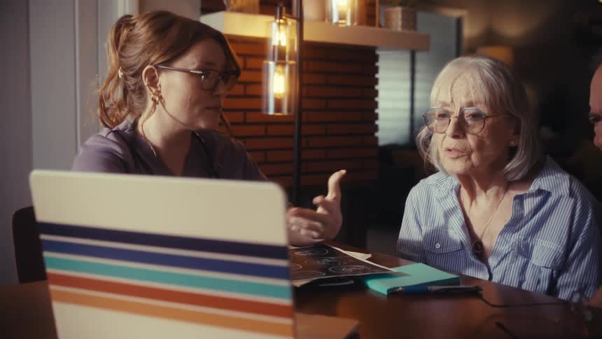 A compassionate doctor explains medical results from a laptop to a worried elderly couple during an in-home consultation
