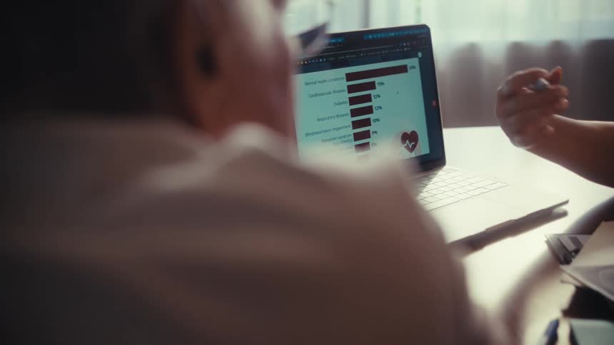 Over the shoulder shot of a woman using a pen to point at a bar graph of medical data on a laptop screen while talking to a senior person