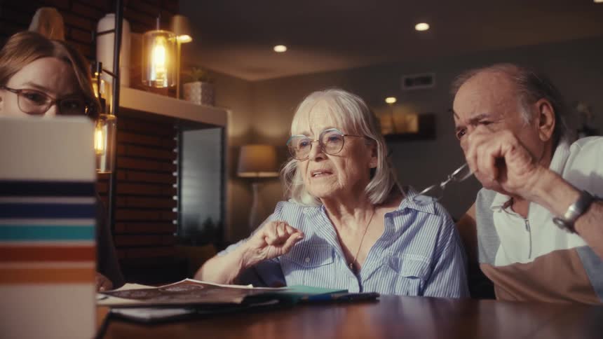 An elderly couple sits with their daughter at a dining room table as she helps them navigate a laptop computer