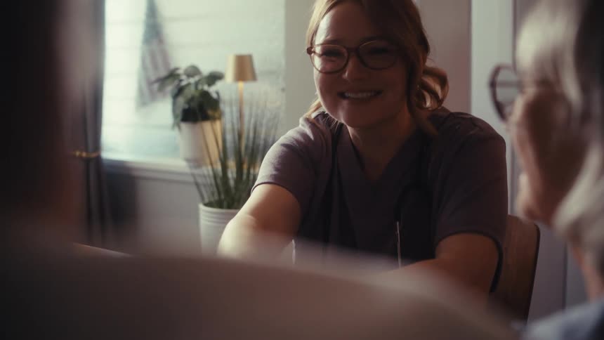 A friendly female doctor smiles and laughs while holding hands with an elderly patient during a house call consultation