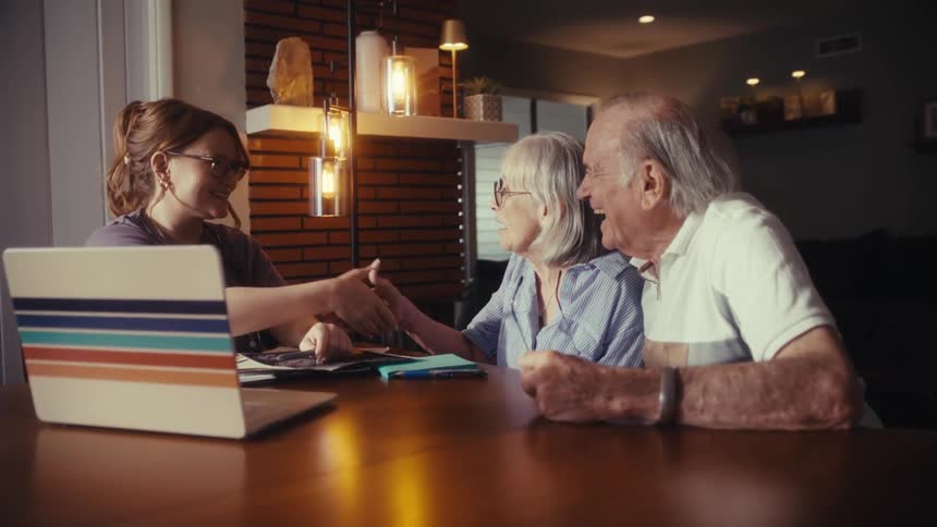 A young female consultant finalizes a deal by shaking hands with a happy elderly couple at their dining table