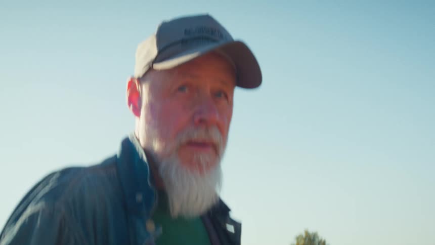 Handheld close-up of an elderly man with a white beard and cap walking outdoors in the sunshine