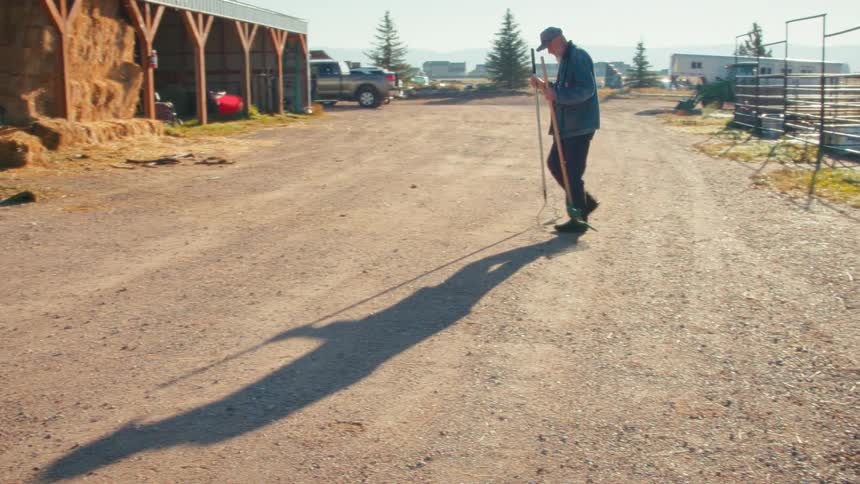 An older farmer walks with a rake across a dirt road on a sunny morning at the ranch, casting a long shadow