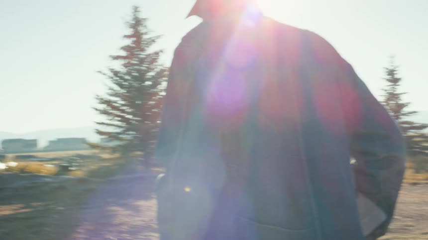 Sun flares over a man in a denim jacket as he pushes a wheelbarrow through a rural landscape