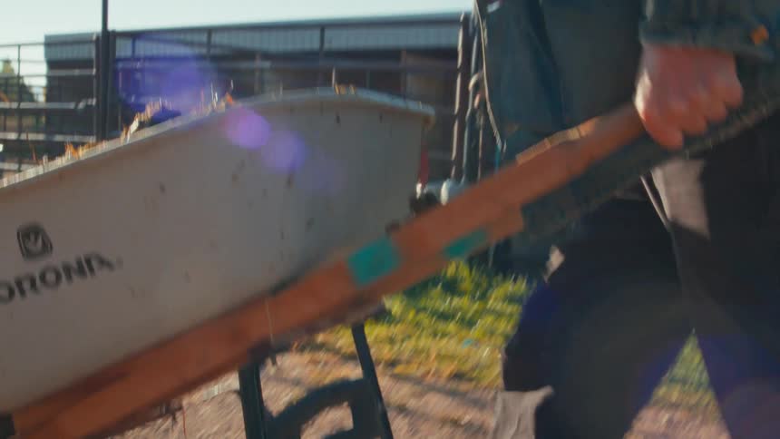 An older farmer with a white beard pushes a wheelbarrow full of steaming manure across a dirt path on the ranch in slow motion